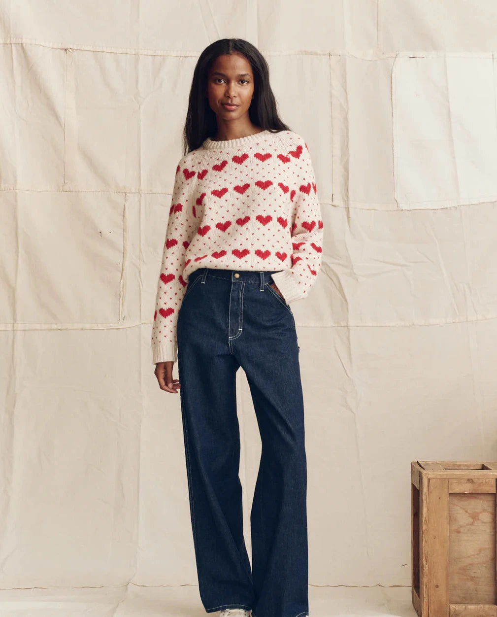 Woman wearing a sweater with heart patterns and jeans standing against a plain wall.